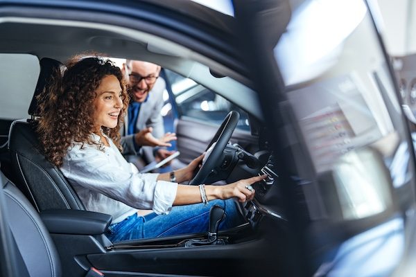 A delighted woman sits in the driver's seat of a car, exploring features, while a salesman enthusiastically assists her in a modern dealership showroom.
