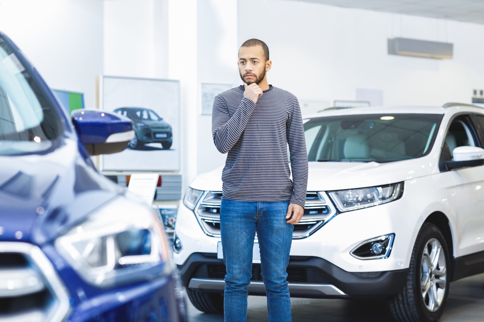 Choice to make. African man choosing a new car at the dealership standing between two vehicles looking confused decision choice buying choosing transport driver buyer consumer client consumerism